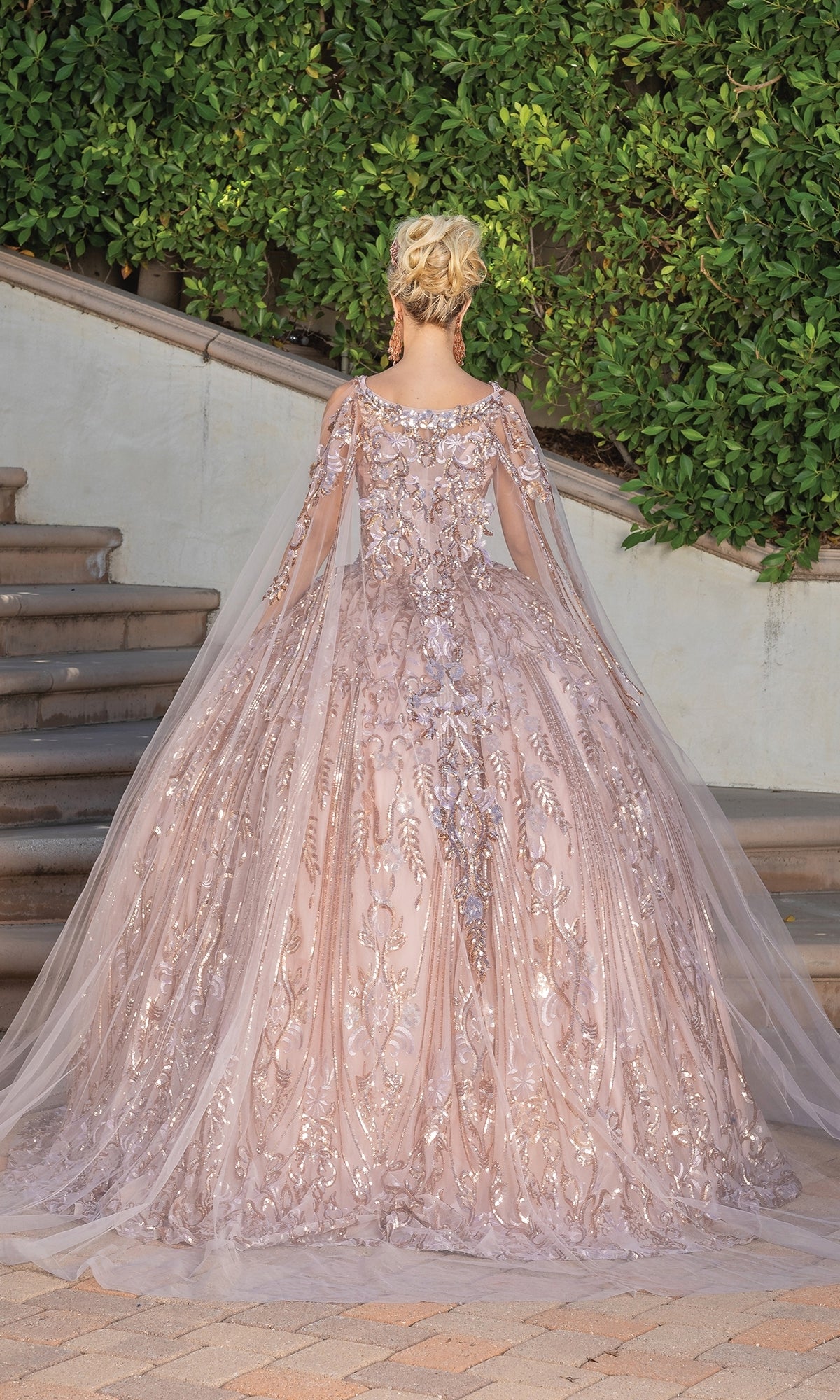 Wearing the Quinceanera Dress 1649 By Dancing Queen—a light pink ball gown with silver details, tulle cape, and sweetheart neckline—a person stands on stone steps, facing away from the camera.
